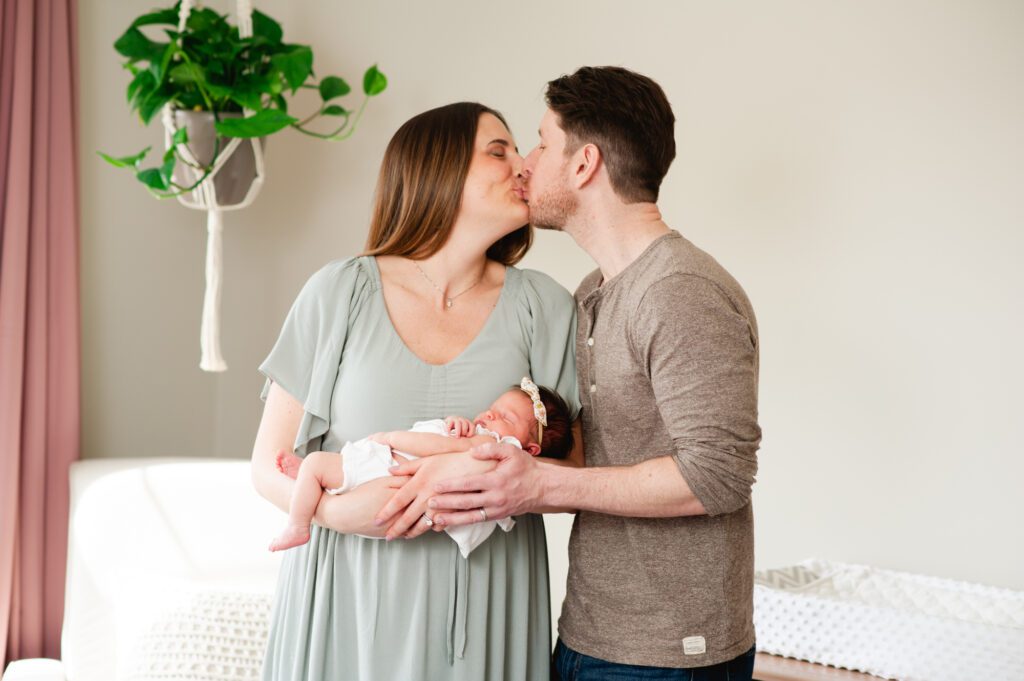 Parents kissing while holding their newborn baby girl in a light-filled nursery