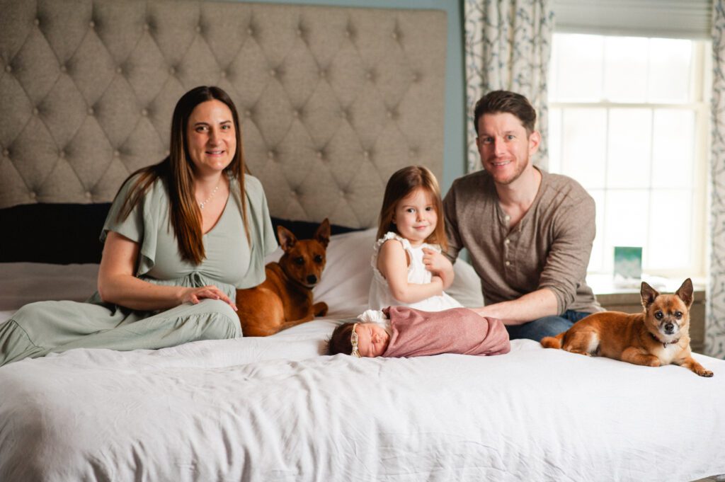 Family smiling and sitting on a white bed with their dogs captured by Philadelphia newborn photographer